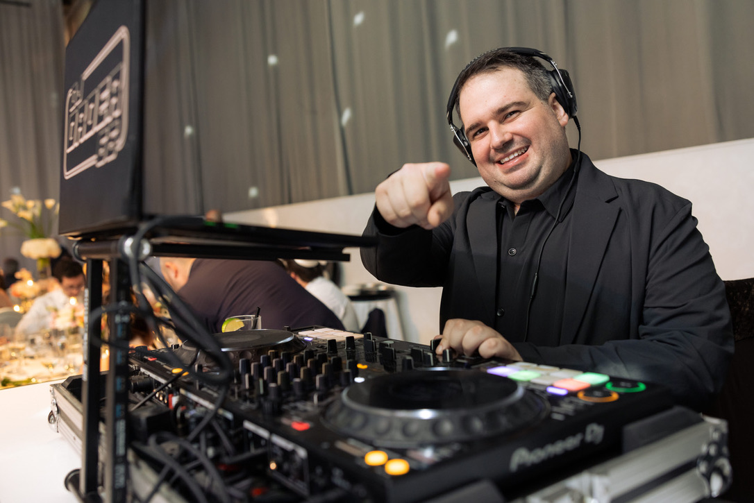 Dylan in a black suit and headphones, pointing at the camera from behind the DJ decks
