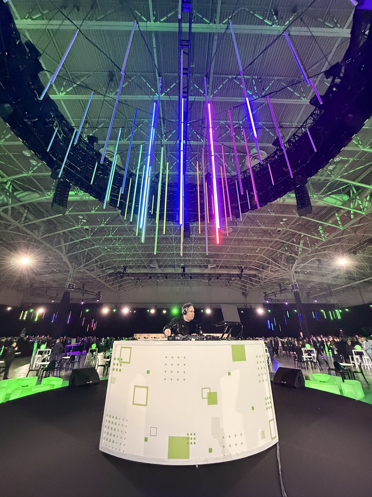 Dylan at a white DJ booth under colourful overhead lighting in a large event venue